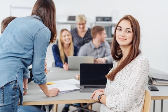 Frau sitzt in einer Gruppe von Mitarbeitenden vor einem Laptop mit Blick in die Kamera