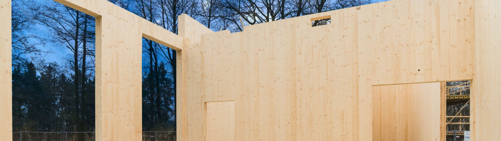 Holzkonstruktion im Bau mit Fenstern und Türen, Betonboden, umgeben von Bäumen und Gerüst.