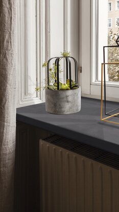 Fensterbank mit modernem Beton-Blumentopf, filigraner Metallverzierung und geometrischer Deko, vor weißem Fenster.