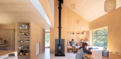 Eine geräumige, lichtdurchflutete Wohnküche, errichtet mit einem Holzwerkstoff-Bausystem, mit einem Kachelofen in der Mitte und drei gemütlich sitzenden Teenagern | © Gayet Roger Architects, FR
