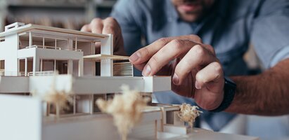 Eine Architekt beugt sich über ein detailliertes Architekturmodell und arbeitet konzentriert in einem hellen, modernen Büro – Symbolbild für kreative Planung und architektonisches Design.