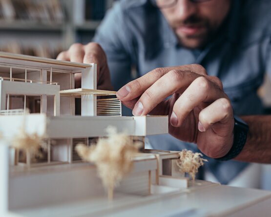 Eine Architekt beugt sich über ein detailliertes Architekturmodell und arbeitet konzentriert in einem hellen, modernen Büro – Symbolbild für kreative Planung und architektonisches Design.