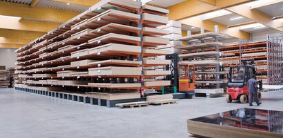 Ein großes Lager mit hohen Regalen, die mit Holzplatten bestückt sind. Die Halle ist hell beleuchtet und hat eine hohe Decke mit Holzträgern.