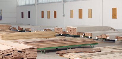 Mehrere Pakete mit dünnen Holzfurnieren liegen gestapelt auf Paletten in einer Lagerhalle. Die Furniere sind aus hellem Holz und teilweise mit Etiketten versehen. Das Bild zeigt die Lagerung von Furniermaterial.