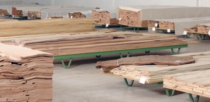 Mehrere Pakete mit dünnen Holzfurnieren liegen gestapelt auf Paletten in einer Lagerhalle. Die Furniere sind aus hellem Holz und teilweise mit Etiketten versehen. Das Bild zeigt die Lagerung von Furniermaterial.