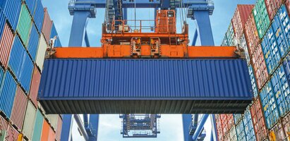 Ein blauer Frachtcontainer wird mit einem Kran gehoben. Im Hintergrund stehen weitere Container. Die Szene zeigt den Umschlag von Waren im Logistik- oder Streckengeschäft.