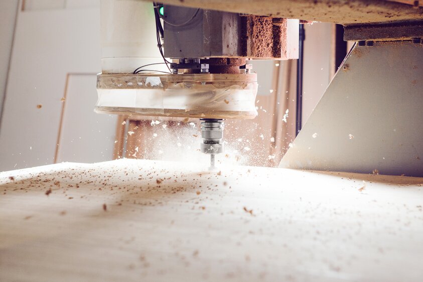 Eine CNC-Fräsmaschine bearbeitet eine rechteckige Holzplatte. Der Fräskopf ist im Einsatz, und Holzspäne fliegen durch die Luft. 
