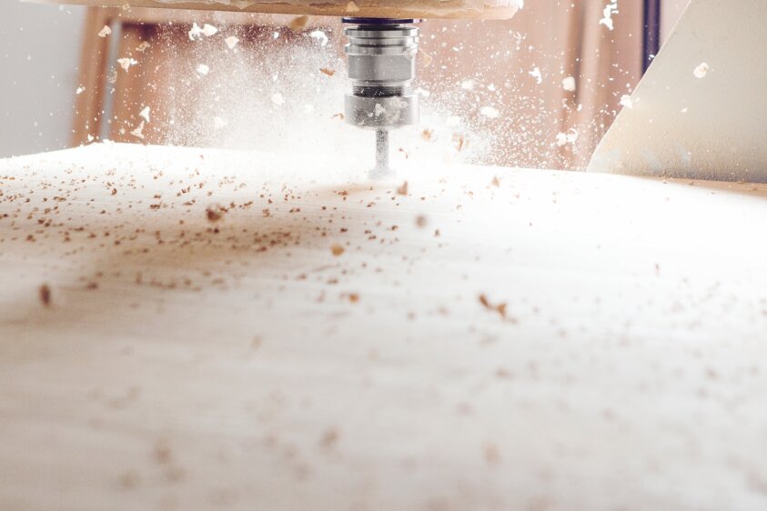 CNC-Fräsmaschine bearbeitet Holzplatte Eine CNC-Fräsmaschine bearbeitet eine rechteckige Holzplatte. Der Fräskopf ist im Einsatz, und Holzspäne fliegen durch die Luft.