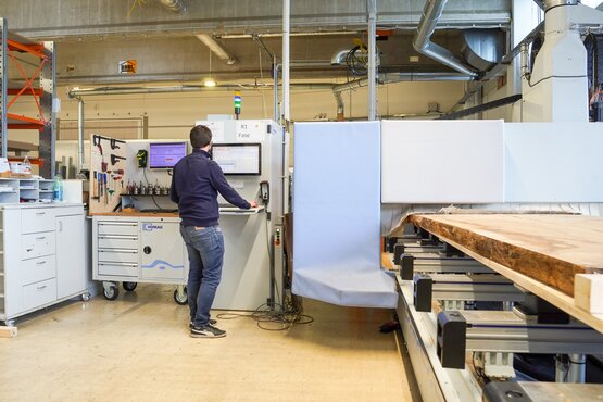 Eine Personsteht vor einer CNC-Maschine und beobachtet die Bearbeitung eines Holzbauteils. Das Werkstück liegt auf einem Maschinentisch. Die Szene zeigt eine manuelle Überwachung in der computergesteuerten Holzbearbeitung.