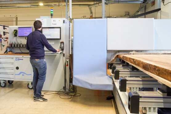 Person bedient Maschine zur Holzbearbeitung Eine Personsteht vor einer CNC-Maschine und beobachtet die Bearbeitung eines Holzbauteils. Das Werkstück liegt auf einem Maschinentisch. Die Szene zeigt eine manuelle Überwachung in der computergesteuerten Holzbearbeitung.