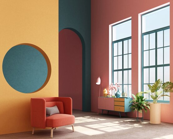 Das Bild zeigt einen hellen, farbenfrohen Raum mit modernen Möbeln in verschiedenen Farben. Die Gestaltung kombiniert warme Holztöne mit kräftigen Farben wie Gelb, Blau und Rosa. Der Raum wirkt freundlich, kreativ und modern