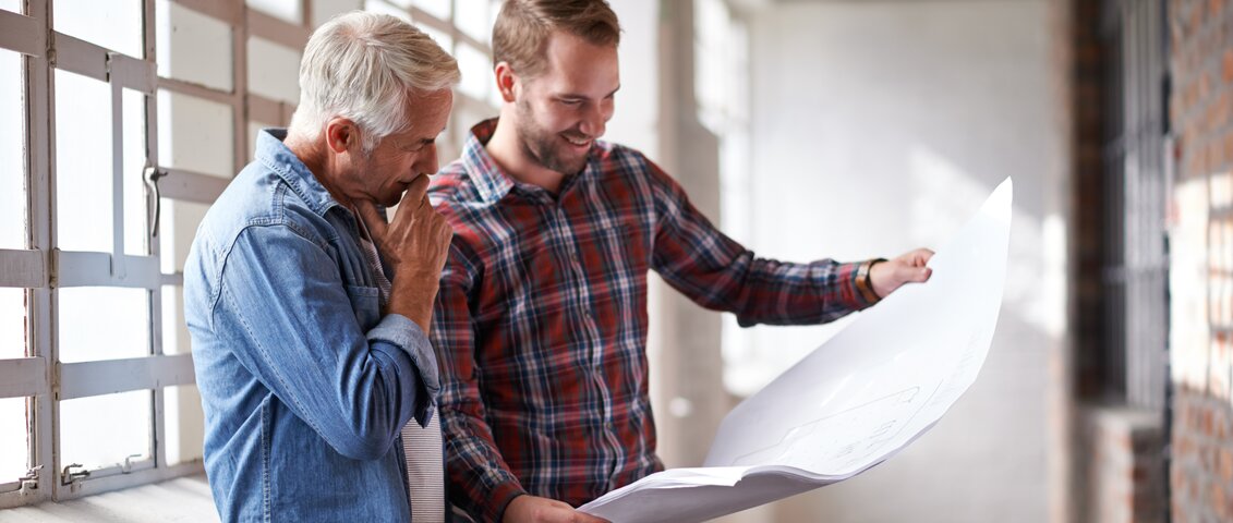 Zwei Männer stehen und betrachten gemeinsam einen großformatigen Plan. Die Szene zeigt eine professionelle Beratungssituation mit Fokus auf Planung, Zusammenarbeit und Projektentwicklung.