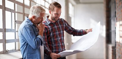 Zwei Männer stehen und betrachten gemeinsam einen großformatigen Plan. Die Szene zeigt eine professionelle Beratungssituation mit Fokus auf Planung, Zusammenarbeit und Projektentwicklung.