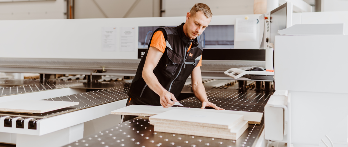 Ein Mann steht an einer Arbeitsmaschine und klebt ein kleines Etikett auf eine zugeschnittene Holzplatte. Die Szene spielt sich in einer modernen Werkstattumgebung ab und zeigt einen Arbeitsschritt im Rahmen der Qualitätskontrolle oder Kennzeichnung im Bearbeitungsservice von JAF.
