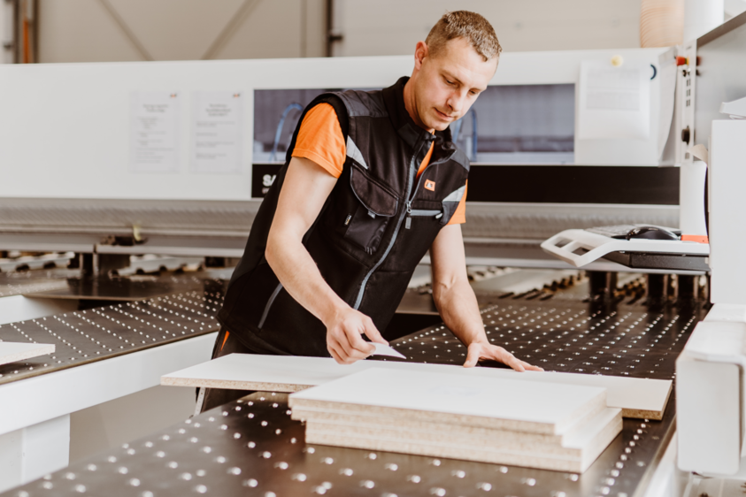 Ein Mann steht an einer Arbeitsmaschine und klebt ein kleines Etikett auf eine zugeschnittene Holzplatte. Die Szene spielt sich in einer modernen Werkstattumgebung ab und zeigt einen Arbeitsschritt im Rahmen der Qualitätskontrolle oder Kennzeichnung im Bearbeitungsservice von JAF.