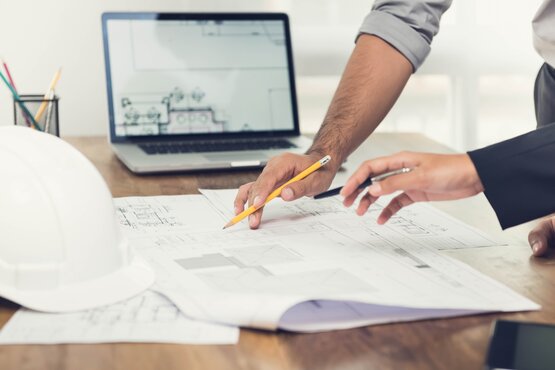 Zwei Personen zeigen mit Stiften auf einen Bauplan Zwei Hände, jeweils mit einem Stift, deuten auf einen ausgebreiteten Plan oder eine technische Zeichnung. Die Szene vermittelt eine konzentrierte Besprechungssituation im Rahmen eines gewerblichen Projekts, bei der Details gemeinsam analysiert oder abgestimmt werden.