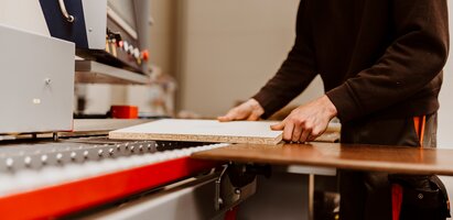 Ein Mitarbeiter bedient eine Maschine, die eine Holz- oder Dekorplatte präzise zuschneidet. Die Platte liegt flach auf dem Maschinentisch. Die Szene zeigt einen professionellen Bearbeitungsprozess im Plattenzuschnitt.
