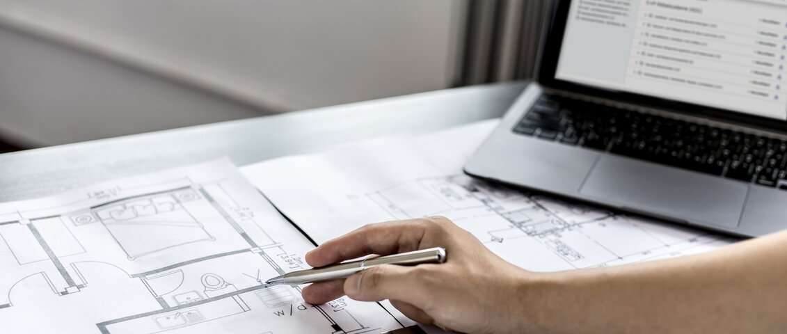 Eine Hand hält einen Stift und zeigt auf einen detaillierten Architekturplan, der auf einem Tisch liegt. Der Plan zeigt Grundrisse mit Linien, Maßen und Beschriftungen. 