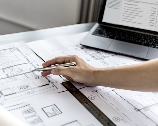 Eine Hand hält einen Stift und zeigt auf einen detaillierten Architekturplan, der auf einem Tisch liegt. Der Plan zeigt Grundrisse mit Linien, Maßen und Beschriftungen. 