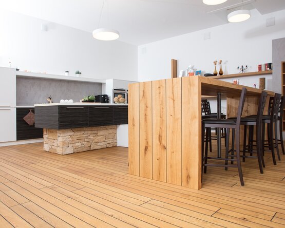 Ein stilvoll eingerichteter Wohnraum mit hellem Holzfußboden in natürlicher Optik. Im Vordergrund steht ein Esstisch mit schwarzen Stühlen, im Hintergrund eine moderne Küche. Große Fenster lassen viel Tageslicht herein und schaffen eine freundliche, offene Atmosphäre. Der Bodenbelag wirkt warm und einladend.