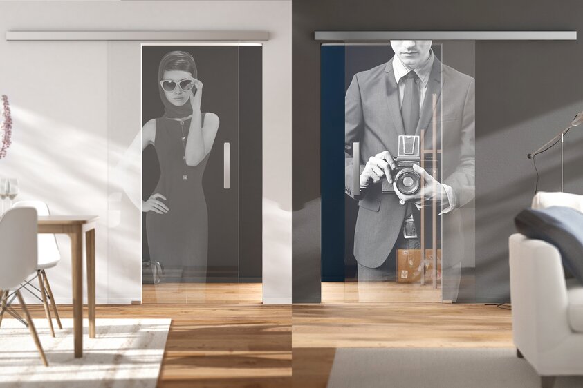 Moderne Glastür „Puristo Planeo“ von Griffwerk in stilvollem Wohnraum Glastür mit Bildern darauf, eingebaut in eine weiße Wand. Links ein heller Sessel mit Beistelltisch, rechts ein graues Sofa. Holzfußboden, helle, moderne Einrichtung