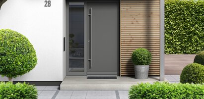 Eingangstür in dunklem Farbton mit vertikalem Glaseinsatz. Die Tür ist in eine helle Fassade und einer Holzverkleidung eingebaut, davor ein überdachter Eingangsbereich mit Pflastersteinen. Links der Tür befindet sich ein schmales, seitliches Glaselemente. Die Szene wirkt modern, einladend und hochwertig.