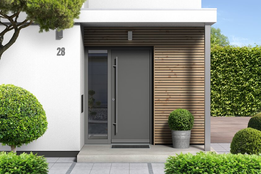 Moderne Hauseingangstür mit Glaselement Eingangstür in dunklem Farbton mit vertikalem Glaseinsatz. Die Tür ist in eine helle Fassade und einer Holzverkleidung eingebaut, davor ein überdachter Eingangsbereich mit Pflastersteinen. Links der Tür befindet sich ein schmales, seitliches Glaselemente. Die Szene wirkt modern, einladend und hochwertig.