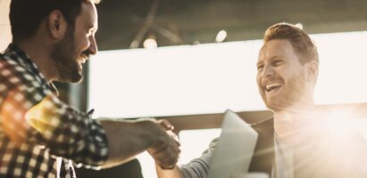 Zwei Männer geben sich die Hand in einer Lagerhalle. Die Sonne scheint tiefstehend und hell durch ein großes Fenster direkt in die Kamera, wodurch ein warmer Lichtschein entsteht. Die Szene vermittelt einen professionellen und zugleich freundlichen Moment der Begrüßung oder Einigung.