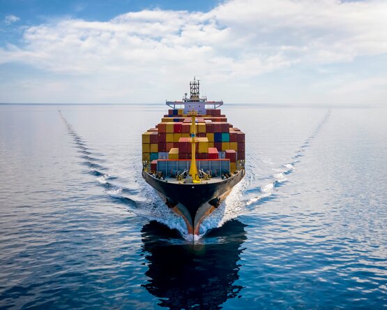 Ein großes Frachtschiff fährt über das ruhige Meer. Es ist mit zahlreichen bunten Containern beladen, die ordentlich gestapelt sind. Der Himmel ist teilweise bewölkt.