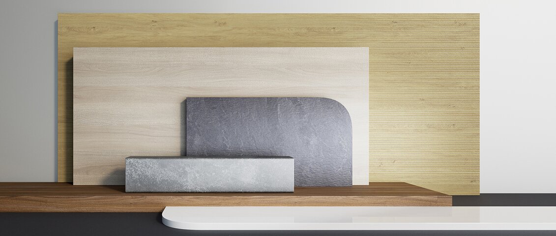 Eine Komposition aus verschiedenen rechteckigen und geometrischen Formen aus unterschiedlichen Materialien. Im Hintergrund befindet sich eine große helle Holzplatte, davor eine mittelgroße beige Holzplatte. Rechts liegt eine kleinere, dunkelgraue Platte mit abgerundeten Ecken, links unten ein länglicher grauer Steinblock. Am Boden liegt eine weitere Holzplanke. Die Szene ist vor neutralem Hintergrund mit weichem Licht arrangiert.