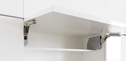 Das Bild zeigt einen geöffneten Küchenschrank mit einem weißen Regalbrett. Sichtbare Scharniere befinden sich an den Seiten. Das Design ist modern und schlicht.