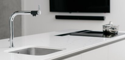 Das Bild zeigt eine moderne Küchenarbeitsplatte mit eingebauter Spüle und Wasserhahn links sowie einem Induktionskochfeld mit Topf rechts. Im Hintergrund sind ein Wandfernseher und eine Soundbar zu sehen. Das Design ist minimalistisch.