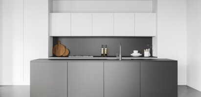 Das Bild zeigt eine moderne Küche mit weißen Oberschränken, grauen Unterschränken, schwarzer Arbeitsplatte und Rückwand. Auf der Arbeitsfläche stehen zwei Holzschneidebretter, zwei Flaschen, Geschirr sowie eine Spüle mit Wasserhahn.