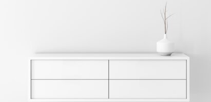 Das Bild zeigt eine weiße Kommode mit vier Schubladen. Auf der Kommode steht eine weiße Vase mit dünnen Zweigen. Der Hintergrund ist schlicht weiß und unterstreicht das minimalistische Design.