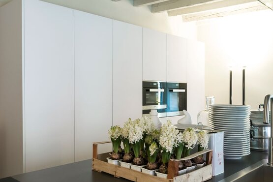 Moderne Küche mit weißen Schränken und Blumenkiste | © Fenix NTM Compact | Nero Ingo | 0720 Das Bild zeigt eine moderne Küche mit weißen Schränken und einem eingebauten Doppelbackofen. Auf der Arbeitsplatte steht eine Holzkiste mit weißen Blumen, daneben ein Stapel weißer Teller und Küchengeräte. | © Fenix NTM Compact | Nero Ingo | 0720