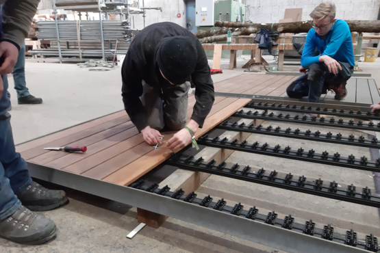 Terrassenschulung mit Holzmaterialien Mehrere Personen nehmen an einer Terrassenschulung im Freien teil. Sie stehen um eine Präsentationsfläche mit Holzmaterialien, während einer daran arbeitet.