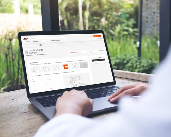 Das Bild zeigt eine Person im Freien, die einen Laptop benutzt. Auf dem Bildschirm ist die Webseite „JAF TERRASSEN-DESIGNER“ mit Gestaltungselementen, Maßen und Diagrammen zur Terrassenplanung zu sehen.