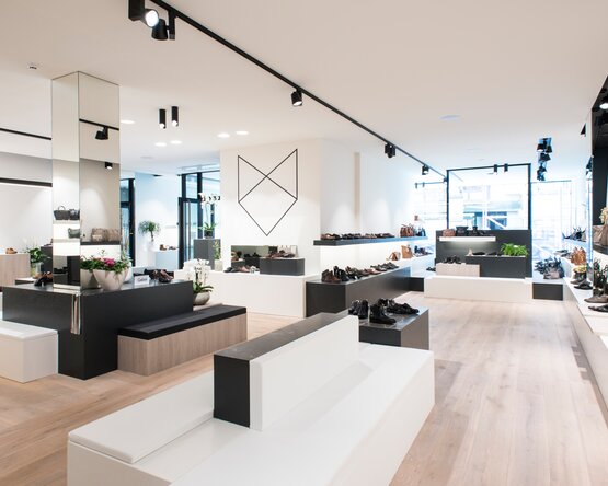 Das Bild zeigt das Innere eines modernen Schuhgeschäfts mit weißen Wänden, hellem Holzboden und schwarzen Akzenten. Auf Regalen sind verschiedene Schuhe ausgestellt, es gibt Sitzgelegenheiten, Spiegel und dekorative Pflanzen. Große Fenster lassen Tageslicht herein.