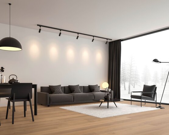 Das Bild zeigt ein modernes Wohnzimmer mit großer Fensterfront und Blick auf eine verschneite Landschaft. Im Raum stehen ein dunkelgraues Sofa mit gemusterten Kissen, ein schwarzer Sessel, ein runder Couchtisch auf weißem Teppich sowie ein Esstisch mit schwarzen Stühlen. Decken- und Stehleuchten sorgen für Beleuchtung.