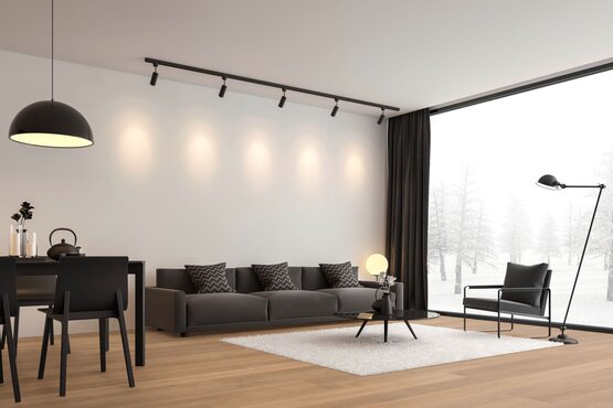 Modernes Wohnzimmer mit Blick auf verschneite Landschaft Das Bild zeigt ein modernes Wohnzimmer mit großer Fensterfront und Blick auf eine verschneite Landschaft. Im Raum stehen ein dunkelgraues Sofa mit gemusterten Kissen, ein schwarzer Sessel, ein runder Couchtisch auf weißem Teppich sowie ein Esstisch mit schwarzen Stühlen. Decken- und Stehleuchten sorgen für Beleuchtung.