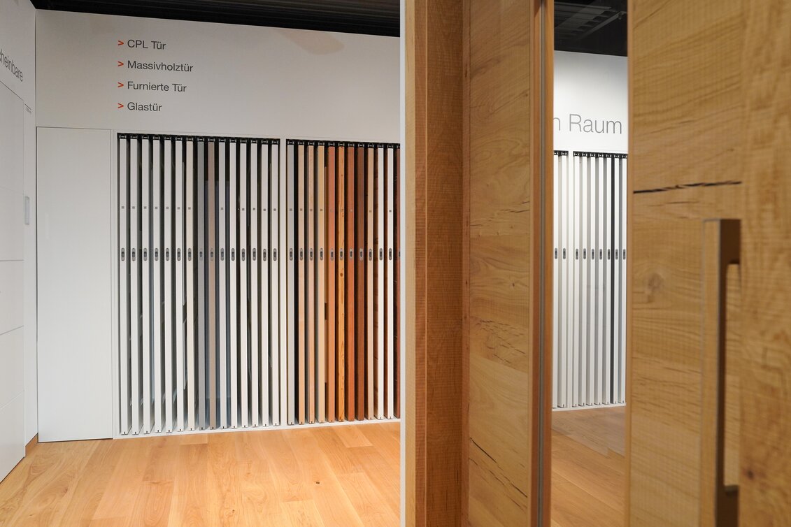 Verschiedene Türmodelle in einem Showroom, mit Kategorien wie CPL-, Massivholz-, furnierte und Glastüren, auf hellem Holzparkettboden.