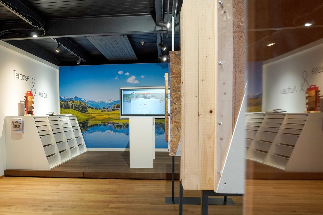 Innenraum mit großem digitalen Bildschirm vor einer Wand mit Berg- und Seepanorama, flankiert von Regalen mit Mustern.