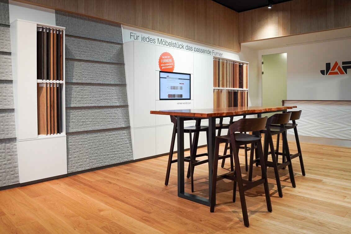 Innenraum mit Holzboden, hohem Tisch und Stühlen, links eine Wand mit Furniermustern und dem Schriftzug „Für jedes Möbelstück das passende Furnier“, daneben ein Bildschirm und im Hintergrund ein „JAF“-Logo.