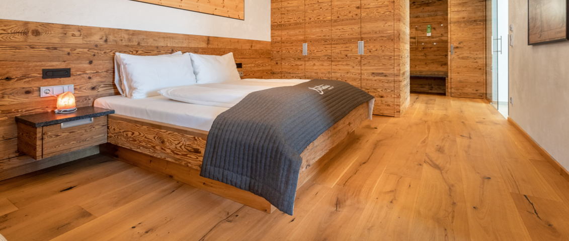 Innenaufnahme eines stilvoll eingerichteten Hotelzimmers. Im Vordergrund ist ein eleganter, dunkler Holzfußboden mit natürlicher Maserung zu sehen. Ein großes, gemütliches Bett mit weißer Bettwäsche steht im Raum, flankiert von modernen Nachttischen. Die Atmosphäre wirkt hochwertig, ruhig und einladend