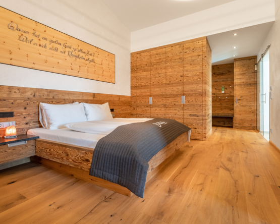 Innenaufnahme eines stilvoll eingerichteten Hotelzimmers. Im Vordergrund ist ein eleganter, dunkler Holzfußboden mit natürlicher Maserung zu sehen. Ein großes, gemütliches Bett mit weißer Bettwäsche steht im Raum, flankiert von modernen Nachttischen. Die Atmosphäre wirkt hochwertig, ruhig und einladend
