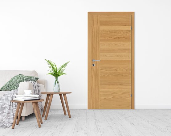 Heller Raum mit Holzfußboden, rechterhand eine helle Holztür, links ein beiges Sofa mit grünen Kissen, davor zwei kleine Holztische mit Pflanze, Büchern und Tasse.