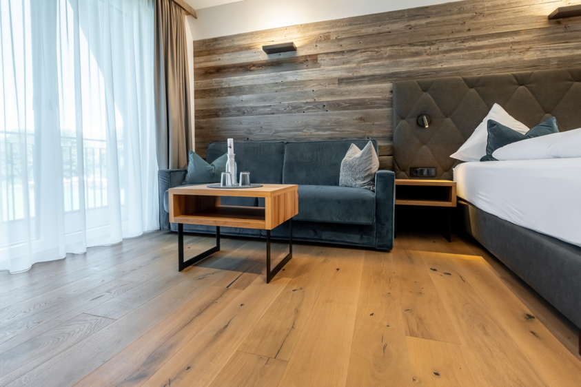 Hotelzimmer im Stubaital mit edlem Bodenbelag aus Holz Innenaufnahme eines stilvollen Hotelzimmers im Hotel Stubai. Der Raum ist mit einem hochwertigen Holzfußboden ausgestattet, der durch seine warme Farbe und natürliche Maserung eine gemütliche Atmosphäre schafft. Im Bild sind ein großes Bett mit heller Bettwäsche, moderne Möbel und eine ruhige, alpine Einrichtung zu sehen.