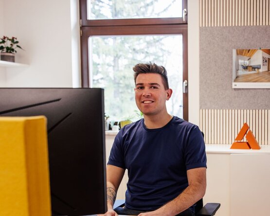 Person sitzt an einem Schreibtisch vor einem Monitor, Gesicht verpixelt; im Hintergrund ein Fenster mit Blick auf Bäume, rechts ein Bildrahmen und ein orangefarbenes Objekt auf einem Regal.