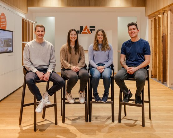 Vier Personen sitzen auf hohen Stühlen in einem Raum mit Holzfußboden. Im Hintergrund ist ein Logo mit den Buchstaben „JAF“ und architektonische Elemente zu sehen.