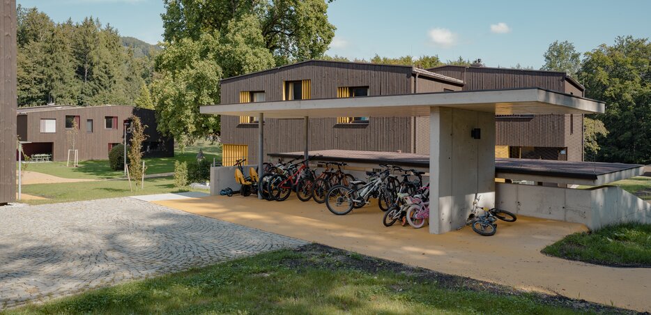 Modernes Gebäude in Holzbauweise mit Flachdach, umgeben von Grünflächen und Fahrradunterstand im Vordergrund. | © LibertydotHome GmbH